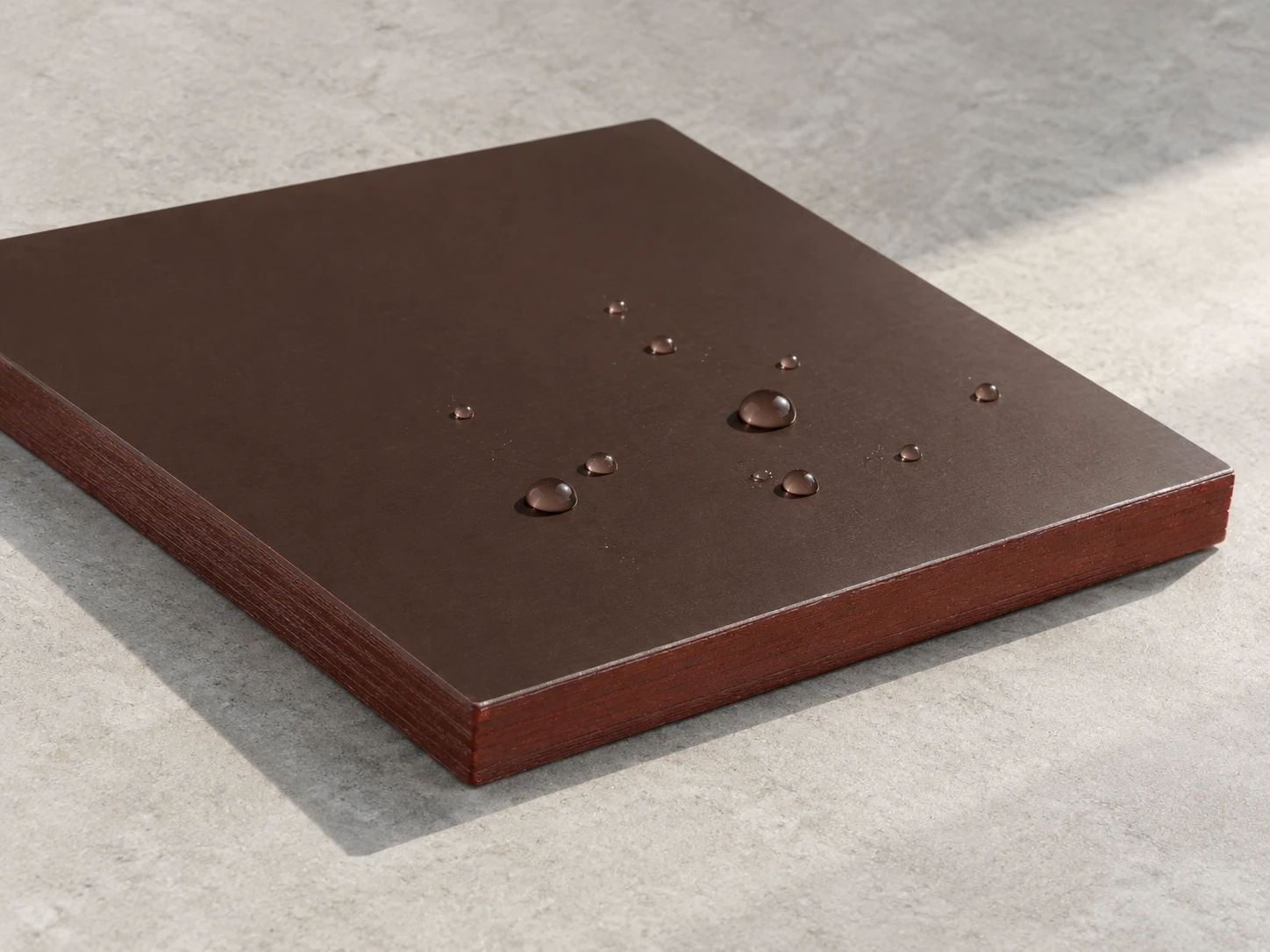 Corner of a dark brown film-faced plywood panel with water droplets beading on the film and a red-brown sealed edge